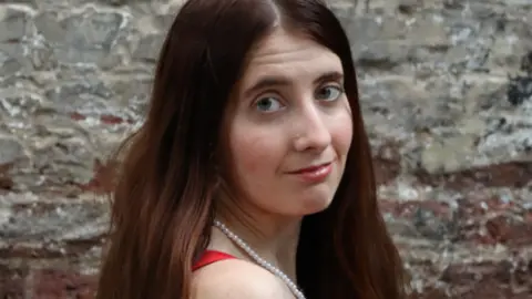 A woman with long brown hair is wearing a white pearl necklace and red strap top. there is a grey stone wall behind her