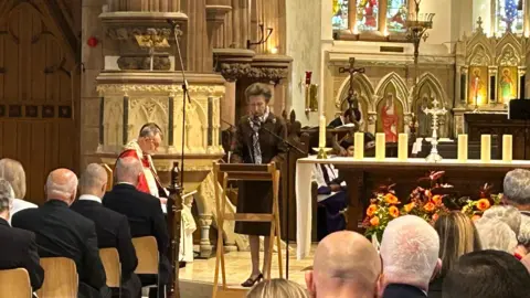 Princess Anne stands and addresses people in Cathedral Isle of Man with extravagant decor in the background. 