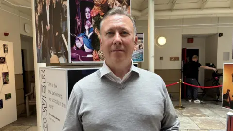 Professor Sean McNamara has grey hair and wears a white shirt with a grey v-neck jumper. He stands in the LIPA building in front of a sign.