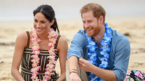 Meghan Markle, wearing a striped black, white and brown dress, sitting next to Prince Harry, wearing a blue collared shirt with sleeves rolled up. Both are smiling and wearing garlands of flowers around their necks. 