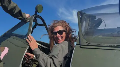 Candy Adkins Candy Adkins sat in the cockpit of a green plane. She is wearing a green top and black sunglasses and is smiling. The sky is blue. 