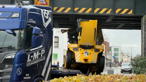 A different view of the stuck lorry and the yellow forklift, looking straight down the street to the buildings and empty road behind the vehicle. A white police van is parked up beyond a line of blue and white police tape