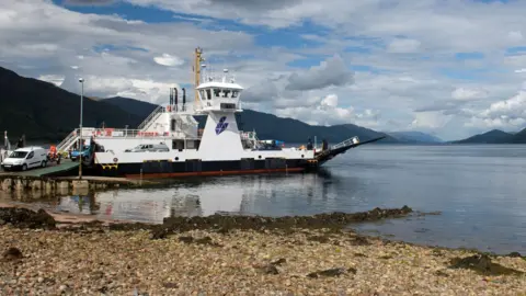 Getty Images The ferry is a white vessel with Highland Council's purple logo just below its bridge. There are vehicles rolling down the boat's green metal ramp on to a slipway. The surface on the loch is calm and there are dark hills along the loch's shores.