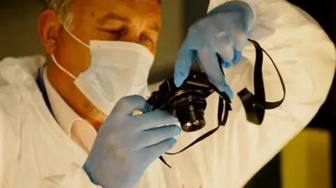 Linsey Smith/ BBC A man wearing a white suit, blue gloves and a facemask holds up a black camera.