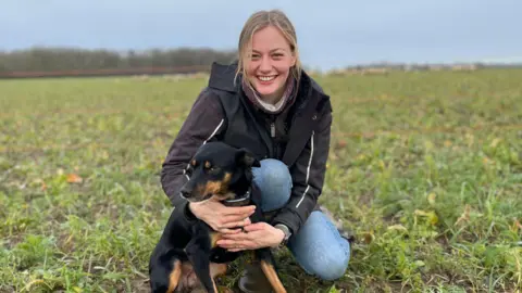 Edd Smith/BBC Hannah Murrell a woman with blonde hair is crouched down in a field with her black and brown sheepdog Roo. She is wearing blue jeans and a black coat. She is looking at the camera and is smiling. 