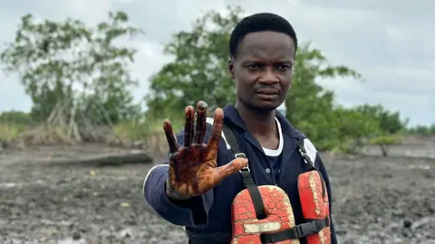 A Nigerian fisherman stood in a naval shirt and stands among an oil-contaminated landscape in Ogoniland, a bathed orange foam life jacket. He puts his hand on the camera - while showing his hand stain with oil in the water.