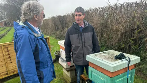 Ian Sexton and Geoff Hardman are standing outside next to two bee hives on a grey day. Ian is looking quizzically at the camera and Geoff is looking at him. Both are wearing anoraks. 