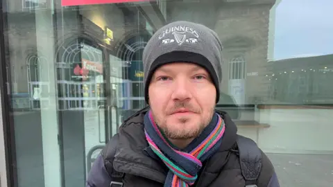 Lee Tyler - a man with a grey beanie, striped scarb and black coat looks directly at the camera