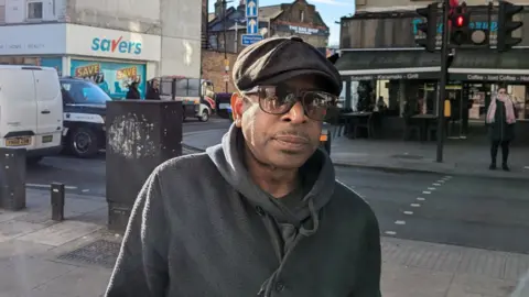 Nev, wearing a grey jacket, glasses and a flatcap, standing on one side of the road with a Savers shop and a cafe on the other side of the road behind him