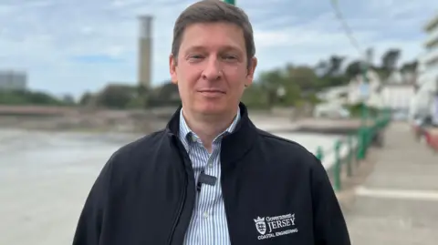 Steve Fitzgibbon standing on the promenade at Havre Des Pas. He has short dark hair. He is wearing a stripey button-up shirt and a "Government of Jersey" zip-up fleece on top.
