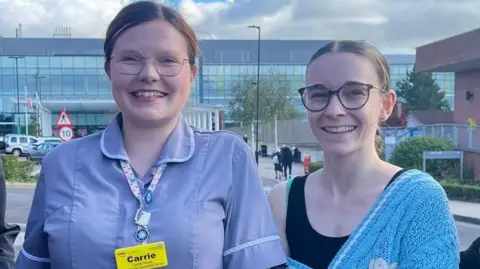 A nurse in a purple uniform with a yellow lanyard with the name Carrie written on it. She has her hair tied back and is smiling at the camera. A woman with blonde hair tied in a plat and glasses is standing next to her, also smiling. The blonde woman is wearing a black vest and a blue cardigan.  The pair are standing in a hospital carpark, with a glass-fronted hospital building and cars seen behind.