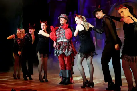 Barry Rivett Hotshots Photography Nigel Harman is in a black and red Fleshcreep costume. His shiny black waistcoat has red braiding and he wears a red shirt underneath with wide sleeves. His top hat is black with red braiding. His hands are on his shoulders in a dance move and he is smiling. He has three dancers on each side of him wearing black costumes with red devil horns.
