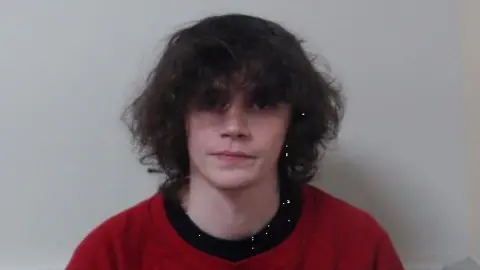 Police Service of Northern Ireland A still of Ezra Garfield. He is sitting in front of a grey wall and is looking at the camera with a neutral expression. He has long, dark hair and is wearing a red t-shirt with a black collar.