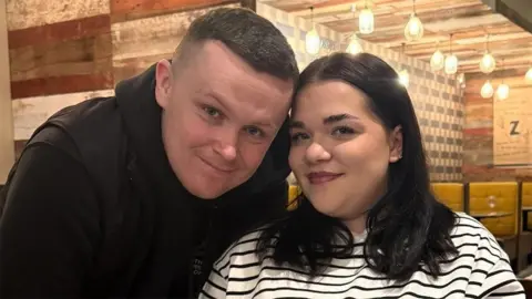Ryan Storey has short black hair and is wearing a black top. He is standing next to his fiancée Anais who has black shoulder-length hair and is wearing a white and blue striped top