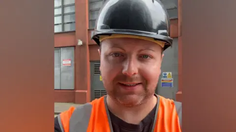 Contributed A man wearing a black hard helmet, orange hi-vis and black T-shirt takes a selfie style image and smiles. He stands in front of a building. 