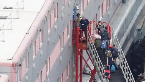 PA Media People carrying bags and suitcases walking up a ramp that leads to the entrance of a grey and red three-storey barge. 