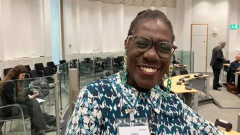 Vikki Irwin/BBC Franstine Jones is wearing a green and white flowered shirt and is standing in the main council chamber at Suffolk's Endeavour house. It has chairs, desks and glass panels. 