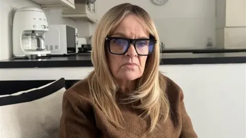 A woman with long blonde hair and glasses and a brown jumper is looking down and looking sad. She is sitting in her kitchen.