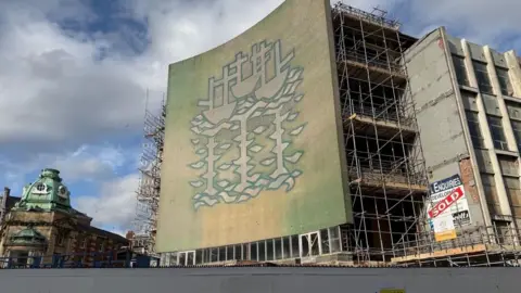 The front of a building with a large concrete mural of three ships. Behind the mural is a partly demolished building with steel and concrete pillars visible.