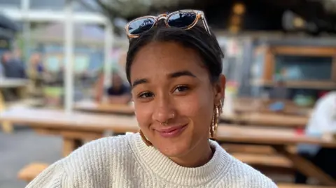 Beth, wearing a white top and with sunglasses perched on her head. She is sitting with some benches behind her which are out of focus