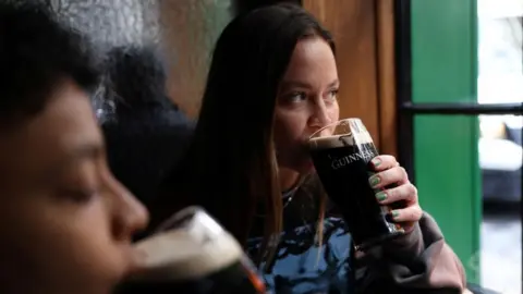 Reuters A woman with dark hair drinks a pint of Guinness beer in a pub while looking out of green-lined window.