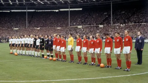 PA Media Two football teams - England in red jerseys and socks, and West Germany in white jerseys and black shorts, line up on a football pitch. the match officials stand in the middle. Crowds can be seen in the stands behind.