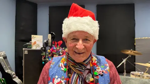Reverend Manville Wiles smiling to camera wearing a Santa hat and festive clothing in recording studio