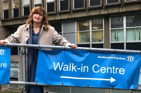 Lucy Galvin Lucy Galvin is wearing a beige overcoat on top of a blue dress. Her hands are resting on a metal railing, from which a blue "Walk-in Centre" sign hangs.