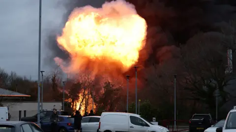 A huge fireball erupts behind some trees. There are orange flames and lots of black smoke. In the foreground there are cars and vans, as members of the public look on.