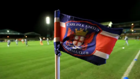 A corner flag at Brunton Park. Football players are training in the background. The flag contains Carlisle United's club shield, which is made up of a red cross, a castle and two dragons.