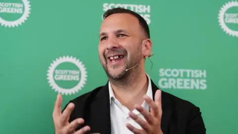 PA Media Zack Polanski, a man with short brown hair wearing a white shirt and black suit jacket. He is mid speech wearing a microphone. He has his hands gesturing in front. Behind him is a green background with the Scottish Greens logo on it.
