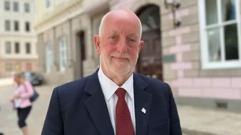 Deputy Steve Luce is standing in front of Jersey's States Assembly building. He is wearing a navy suit with a red tie. He is smiling into the camera.