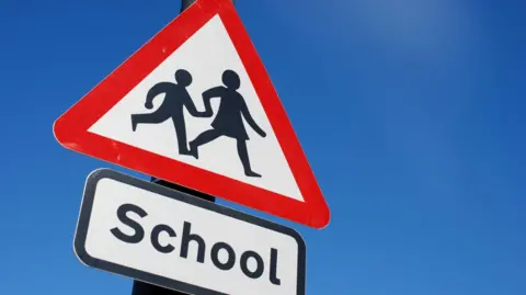 A triangle-shaped sign showing a simple silhouette of a girl holding hands with a smaller boy as they walk. The figures are on a white background with a red border. Underneath the triangle is a rectangle-shaped sign with the word "school".