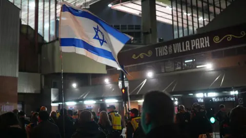 Christopher Furlong/Getty Images An Israeli flag held by pro-Israeli supporters outside Villa Park ahead of the game on 6 November 2025. It is night and police are escorting them