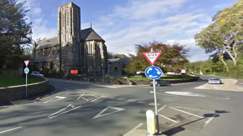 A roundabout next to a large brown brick built church building. There is a give way sign at the roundabout. and trees lining the road on the right.