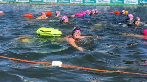 Presenter, Kate Kinsella smiles whilst immersed in river water. She is surrounded by ten other swimmers