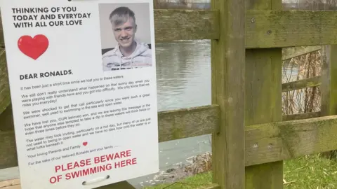 Martin Heath/BBC A wooden fence by a river, with a grass bank visible in the foreground. A sheet of card is fixed to the fence with string. It reads "thinking of you today and every day with all our love, dear Ronalds".  Below further text, there is black writing which says "please beware of swimming here".