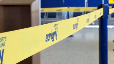 A yellow retractable barrier at Guernsey Airport, which says 'Guernsey's Airline Aurigny'.