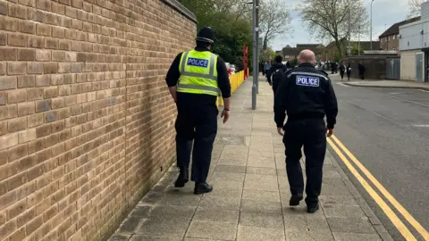 The backs of two police officers walking down a path, with people ahead of them. A brick wall is to the left and double yellow lines on a road to the right. Buildings are to the right and trees ahead. 