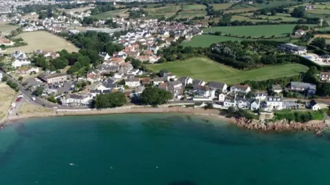 A coastal community of houses along a beach and a beachfront road with fields behind.