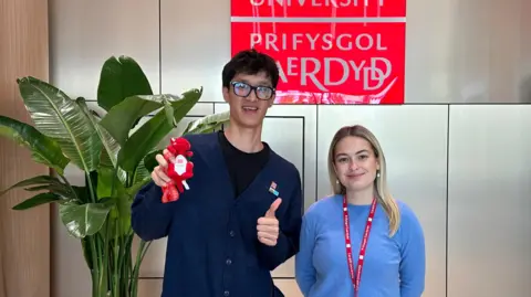 Madison Hutchinson A young man with glasses holding a small red dragon soft toy and with thumbs up, standing next to a young woman with a blue jumper and red lanyard in front of a Cardiff University sign.