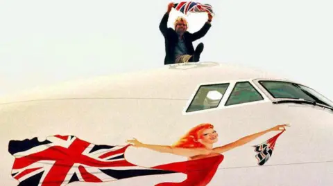 Richard Branson sits on top of a large white plane - only part of the plane can be seen. The windows for the pilots cockpit and underneath where there is the painting of the Scarlet Lady. She is smiling, looking like she is flying forward, wearing a read leotard and holding a billowing union jack flag behind her.