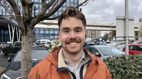 The picture shows a man smiling at the camera. He has brown hair and is wearing an orange jacket. He's standing in front of a carpark, with a building also behind him.