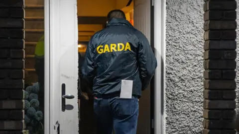 An officer entering a property front door, the officer is wearing a coat that says Garda in yellow. 