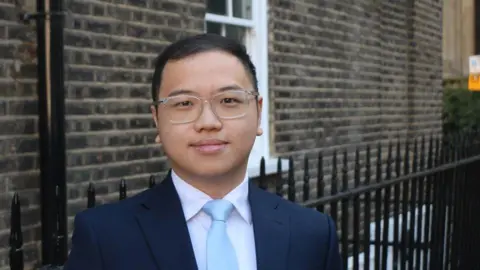 The Taxpayers' Alliance Shimeon Lee with short black hair looking at the camera while wearing clear-framed glasses, a blue jacket, pink shirt and light blue tie. He is standing in front of a brown-bricked building with black railings outside.
