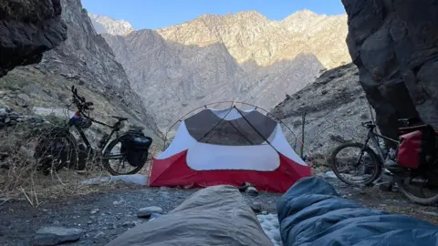 Olly Hargreaves Two bikes are parked either side of a tent, mountains can be seen in the distance, two sleeping bags can be seen in the foreground.
