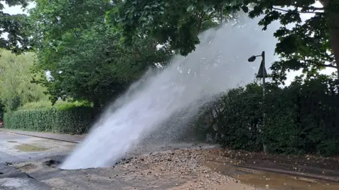 Water shoots out of a large hole in a road - with debris surrounding it.