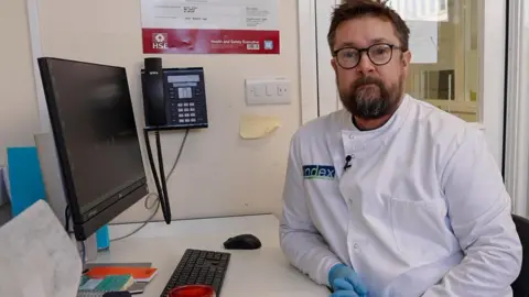 Dr Rob Morley sitting at a white desk with a computer monitor and petri dishes. There is a phone on the wall and anti-bacterial wipes on the desk. Dr Morley has short dark hair and a beard with black glasses. He is wearing a white lab coat and blue PPE gloves.