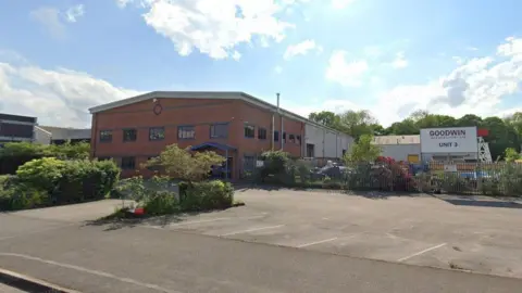 Google A brick-built building on an industrial estate behind metal fencing and a car park. There is a sign that reads Goodwin International Ltd Unit 3.