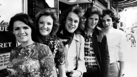 Mirrorpix vía Getty Images Imagen en blanco y negro de Eve Graham (izquierda) con otros miembros de New Seekers en agosto de 1971 (de izquierda a derecha) Lyn Paul, Marty Kristian, Peter Doyle y Paul Layton parados en una calle junto a una tienda. Todos sonríen.